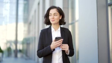 Resmi takım elbiseli genç gülen işadamı ofis binasının yakınındaki cadde boyunca yürürken akıllı telefon kullanıyor. Kadın yönetici mesajlaşıyor, e- postaları kontrol ediyor, çevrimiçi sohbet ediyor veya sosyal medyada geziniyor