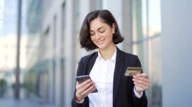 Mutlu genç iş kadını internette alışveriş yapıyor. Bir ofis binasının yanındaki caddede duran akıllı telefondan kredi kartı numarasını yazıyor. Gülümseyen ve tatmin olmuş esmer bir kadın mutlulukla alışveriş yapar.