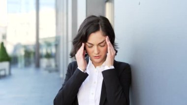Resmi takım elbiseli üzgün genç iş kadını bir ofis binasının yanında dikilirken baş ağrısından muzdarip. Kadın elleriyle şakaklarına masaj yapıyor. Migreni ya da düşük kan basıncı var.