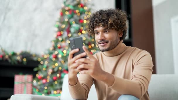 ハッピーな若者は冬の新年のクリスマス休暇中に家でソファーに座っている間 スマートフォンで素晴らしいニュースを受け取りました 興奮したハンサムな男性は電話で楽しいメッセージを読み 成功を祝います — ストック動画