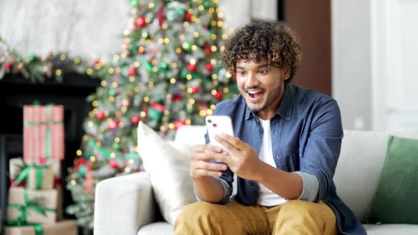 新年佳节期间 快乐的年轻人坐在沙发上 一边用智能手机听到好消息 一个英俊的男人在电话上读到一个愉快的信息 庆祝成功 — 图库视频影像