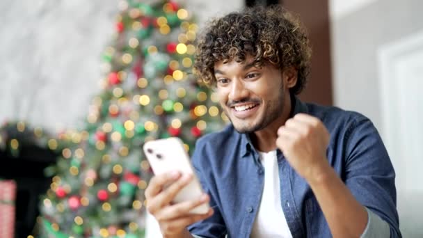 ハッピーな若者は 冬の新年のクリスマス休暇中に家に座っている間 スマートフォンで素晴らしいニュースを受け取りました 興奮したハンサムな男性は 電話で楽しいメッセージを読み 成功を祝います クローズアップ — ストック動画