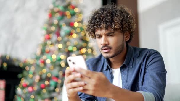 クローズアップ ストレスを受けたハンサムな男性は 冬の新年のクリスマス休暇中に家に座っている間 スマートフォンで悪いニュースを読みます 心配した動揺男性は否定的なメッセージをレビューしながら悲しいです — ストック動画