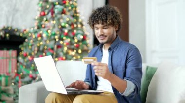Mutlu genç adam, yeni yıl tatilinde evde oturup bilgisayarında kredi kartı numarasını yazarak online alışveriş yapıyor. Gülümseyen erkek ürün tanıtımından memnun kalarak alışveriş yapıyor.