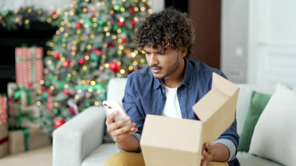 若い男性は 冬の新年のクリスマス休暇中に自宅で間違った製品と小包を受け取った後 スマートフォンで悪いレビューを書いています 失望した男はオンラインストアでショッピングに不満 — ストック動画