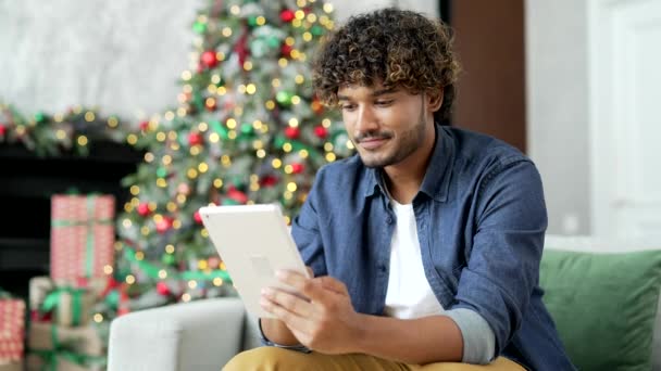 在过年的寒假 一个年轻人坐在家里的沙发上 一边用平板电脑 英俊的男性正在浏览社交网络 写问候语 与朋友聊天或上网购物 — 图库视频影像