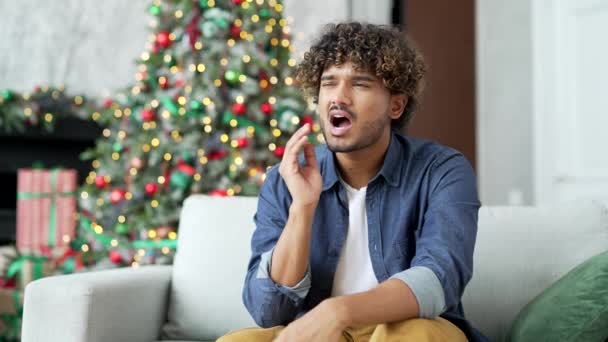 冬の新年のクリスマス休暇中に自宅のリビングルームに座って歯痛に苦しむ若い男 失望した男は頬の近くに手を保持し 痛みの場所をマッサージ — ストック動画