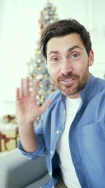 POV Webcam görüntüsü. Dikey video. Yılbaşı Noel tatilinde evde akıllı telefon kullanan mutlu bir adam görüntülü konuşma yapıyor. Gülümseyen sakallı erkek dileklerini uzaktan tebrik ediyor.