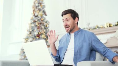 Gülümseyen adam, yılbaşı Noel tatilinde evdeki kanepede oturmuş dizüstü bilgisayarla bir video görüşmesinde sohbet ederken hediye kutusu gösteriyor. Dilek dileyen mutlu adam, uzaktan tebrik ediyor.