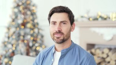 Yılbaşı tatilinde evdeki oturma odasında oturan yakışıklı adamın yakın portresi. Bir Noel ağacının arka planında kameraya bakarak gülümseyen, pozitif sakallı bir erkek resmi.