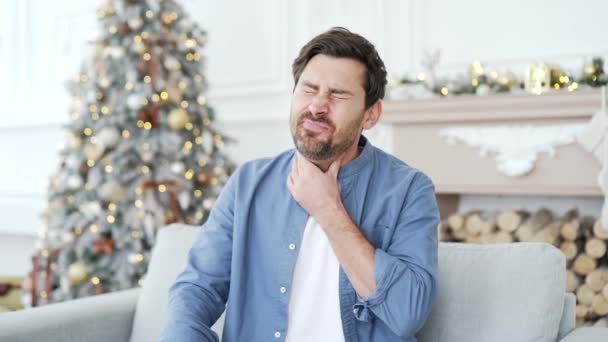 冬のクリスマスXmas休暇中に自宅のリビングルームでソファーに座っている痛みの喉に苦しんでいる病気の男 男性は ウイルスまたはインフルエンザの症状を持っています 痛みのスポットをマッサージ — ストック動画