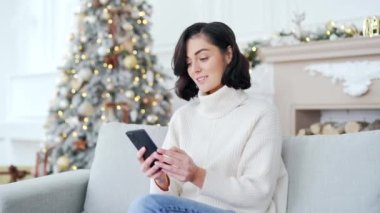 Mutlu, heyecanlı genç kadın, yılbaşı Noel tatilinde evdeki koltukta otururken akıllı telefondan harika haberler aldı. Kız telefonda hoş bir mesaj okuyor, başarıyı kutluyor.