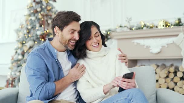 笑顔のカップルは 冬のクリスマスXmas休暇中に家でリビングルームに座っているスマートフォンを使用してオンラインストアで買い物しています 夫と妻は割引と販売に満足しています — ストック動画