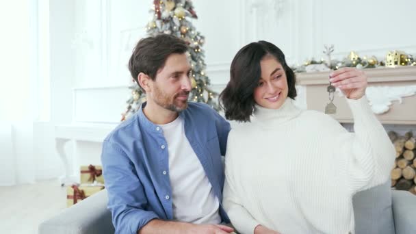 在圣诞佳节期间 一对快乐的新婚夫妇坐在客厅的沙发上 看着摄像机 展示新房子的钥匙 妻子和丈夫拥抱微笑 — 图库视频影像