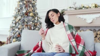 Soğuk algınlığı ya da battaniyeye sarılı hasta bir kadın Noel Noel tatilinde evdeki kanepede oturuyor. Dişi termometreyi elinde tutuyor, ateşi yüksek, çay içiyor.
