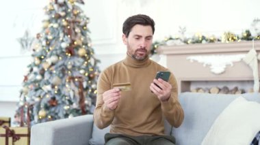 Mutlu bir adam, yılbaşı Noel tatilinde evde oturup akıllı telefondan kredi kartı numarasını yazarak online alışveriş yapıyor. Gülümseyen erkek ürün tanıtımından memnun kalarak alışveriş yapıyor.