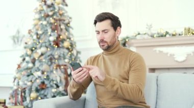 Gülümseyen sakallı bir adam yılbaşı Noel tatilinde evdeki koltukta oturan akıllı telefonu kullanır. Yakışıklı bir erkek arkadaşlarıyla sohbet etmek ya da internetten alışveriş yapmak için bir kutlama mesajı yazıyor.