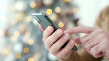 Kapatın. Yılbaşı tatilinde evde akıllı bir telefon tutan bir adam var. Bir erkek, kapalı mekanlarda cep telefonu kullanarak Noel indirimleri, satışları ve tanıtımlarla çevrimiçi bir mağazadan alışveriş yapıyor.