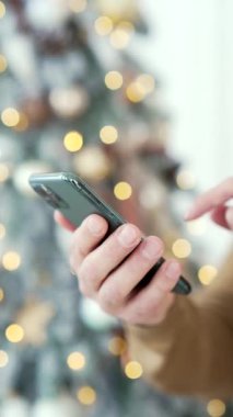 Kapatın. Dikey video. Yılbaşı tatilinde evde akıllı bir telefon tutan bir adam var. Noel indirimleri, satışlar ve promosyonları cep telefonu kullanarak çevrimiçi mağazada erkek alışverişi