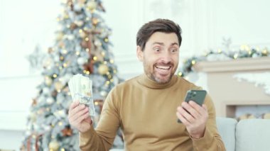 Nakit para ve akıllı telefon hayranı heyecanlı bir adam. Yılbaşı Noel tatilinde evde oturuyor. Erkek, telefonundaki başvuru formunu kullanarak para kazanmaktan memnun.