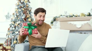 Gülümseyen adam, yılbaşı Noel tatilinde evdeki kanepede oturmuş dizüstü bilgisayarla bir video görüşmesinde sohbet ederken hediye kutusu gösteriyor. Dilek dileyen mutlu adam, uzaktan tebrik ediyor.