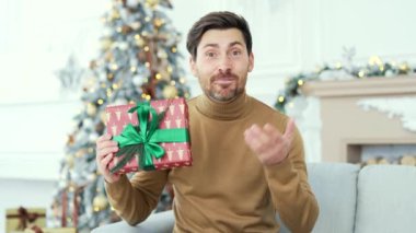 Webcam görüntüsü. Gülümseyen adam, yılbaşı Noel tatilinde evdeki kanepede otururken video görüşmesinde sohbet ederken hediye kutusu gösteriyor. Dilek dileyen mutlu adam, uzaktan tebrik ediyor.