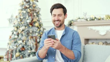 Gülümseyen sakallı bir adam yılbaşı Noel tatilinde evdeki koltukta oturan akıllı telefonu kullanır. Yakışıklı bir erkek arkadaşlarıyla sohbet etmek ya da internetten alışveriş yapmak için bir kutlama mesajı yazıyor.