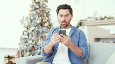 Hüsrana uğramış stresli adam yılbaşı Noel tatilinde evde oturup akıllı telefondan kötü haberler okuyor. Endişeli erkek, cep telefonundaki olumsuz mesajı incelerken üzgün