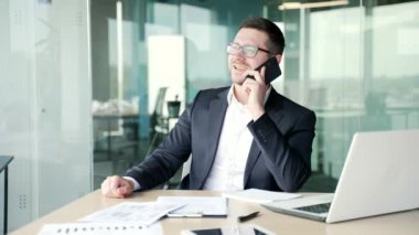 İş yerinde otururken akıllı telefondan konuşan kendine güvenen güler yüzlü iş adamı. Resmi takım elbiseli ve gözlüklü girişimci müşterisiyle iletişim kurar, iş görüşmesi yapar.