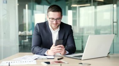 Resmi takım elbiseli mutlu iş adamı iş yerinde otururken akıllı telefondan harika haberler aldı. Neşeli girişimci hoş bir mesaj okur, başarıyı kutlar evet jestleri gösterir