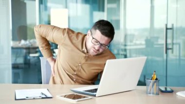 İş yerindeki çalışma masasında dizüstü bilgisayarla çalışırken sırt ağrısı çeken yorgun genç bir işadamı. İşçi masajı sırt kaslarını ağrıtır. Çok yorgun ve çok çalışıyor.