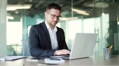 İş yerindeki bir iş yerinde otururken dizüstü bilgisayarda yazı yazan resmi takım elbiseli bir iş adamı. Gözlüklü bir girişimci bilgisayarda bir proje üzerinde çalışıyor ya da internette sohbet ediyor.
