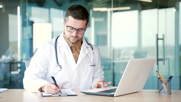 一位穿着白色外套的自信的医生正在医院诊所的笔记本电脑上打字 并在笔记本上写下数据 男医生填写表格或开药方 — 图库视频影像