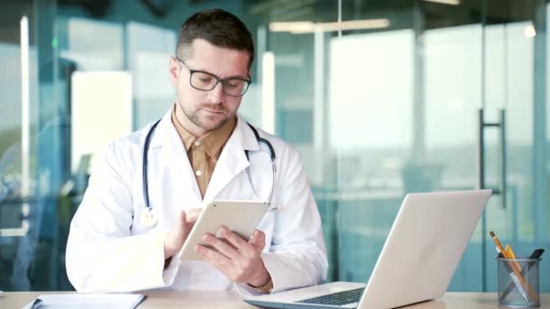 坐在现代医院诊所的工作场所时 身穿白色外套的自信的医生使用数字平板电脑 戴眼镜的医护人员在网上与病人聊天或上网浏览新闻 — 图库视频影像