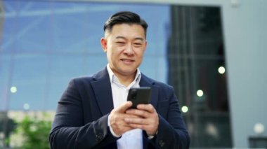 Resmi takım elbiseli gülümseyen iş adamı, bir ofis binasının yanında caddede dururken akıllı telefon kullanıyor. Mutlu girişimci telefonunu karıştırıyor, e-posta mesajlarını kontrol ediyor, internette sohbet ediyor, mesajlaşıyor.