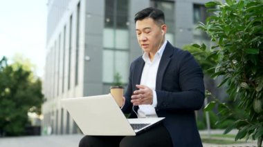 Resmi takım elbiseli Asyalı bir işadamı, ofis binasının yakınındaki bir bankta dizüstü bilgisayar kullanarak bir video görüşmesinde konuşuyor. Girişimci elinde kahve tutarken internetten iletişim kuruyor.