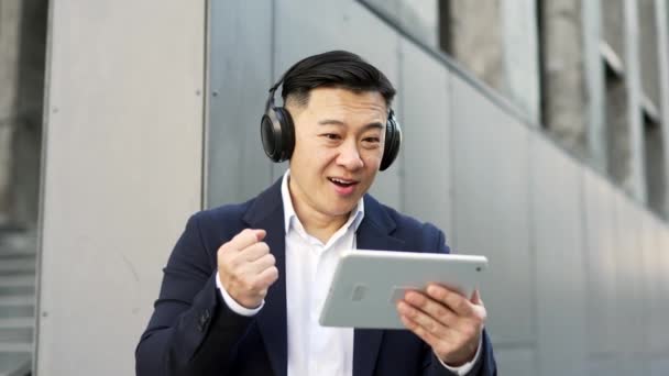 Podekscytowany Biznesmen Azjatycki Formalnym Garniturze Słuchawkach Bezprzewodowych Oglądając Mecz Sportowy — Wideo stockowe