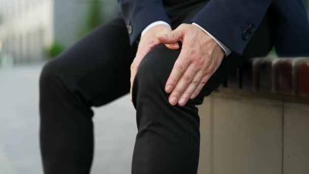 ストリートのベンチに座っている間 膝の筋肉をマッサージする男性の手を閉じます 正式なスーツの男性は 痛みを伴うひざ関節痛に苦しんでおり 歩くことができず クランプ リウマチ — ストック動画