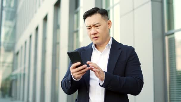 身穿正装的忧心忡忡的亚洲商人站在一幢办公楼附近的街上 在智能手机上看到了坏消息 沮丧的有压力的创业者在电话里看到信息 心情很不好 — 图库视频影像