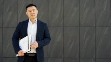 Resmi takım elbiseli mutlu bir Asyalı işadamının portresi ofis binasının karanlık duvarının yanında duruyordu. Dizüstü bilgisayarı ve klasörüyle kameraya bakan gülümseyen bir erkek. Reklam şablonu. Boşluğu kopyala