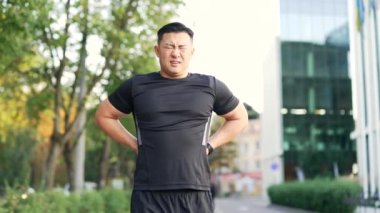 Yetişkin Asyalı koşucu sırt ağrısı, spor sakatlığı, sokak ortasında kas spazmı geçiriyor. Spor kıyafetli bir adam, şehir parkında dışarıda koşudan sonra sırt ağrısı ve sorun yaşıyor.