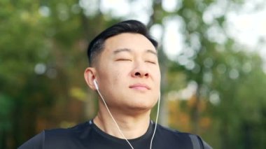 Şehir parkında dikilen mutlu Asyalı sporcunun sabah koşusundan sonra gözleri kapalı dinlenmesi. Kulaklıklı sakin erkek derin nefes alır ve dışarıda müzik dinlemekten hoşlanır.
