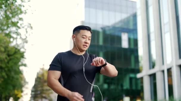 アジアのアスリートランナーは 街路の建物の近代的な都市背景を走っています フィットネストラッカーブレスレットやスマートウォッチの結果を見て屋外ジョギングヘッドフォンでスポーティな男ジョギング — ストック動画