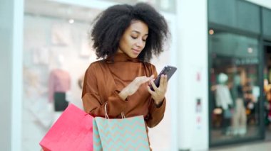 Genç, güzel, mutlu, alışveriş merkezinin alışveriş merkezinde neşeli müşteri duruşu yapıyor ve cep telefonuyla sohbet ediyor, daktilo ediyor akıllı telefon kullanıyor mağazadaki hediye renk paketinden memnun.
