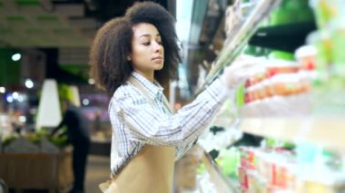 Hipermarkette çalışan Afrikalı Amerikalı çalışan tezgahtar, marketin yanındaki raflarda ürünün durumunu kontrol ediyor. Siyah kadın, yönetici süpermarketteki son kullanma tarihini kontrol ediyor.