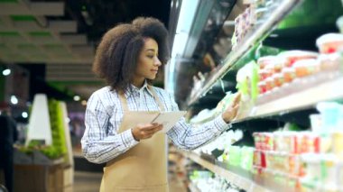 Tablet üzerinde çalışan Afrikalı Amerikalı çalışan tezgahtar, rafların yakınındaki bir markette ürünlerin durumunu kontrol ediyor. Siyah kadın, yönetici süpermarketteki son kullanma tarihini kontrol ediyor.