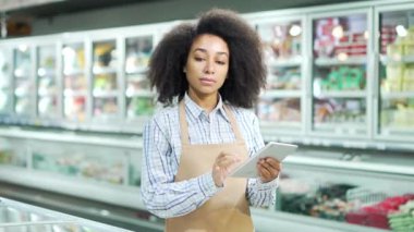 Önlüklü kadın satış elemanı portresi süpermarketteki kameraya bakıyor. Afrikalı Amerikalı siyahi kadın işçi ya da küçük işletme sahibi. İç mekan. Yiyecek, market alışverişi gülümsemesi.