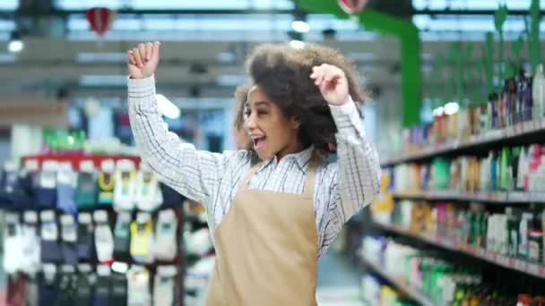 愉快的超级市场女售货员在市场上跳舞 快乐有趣的黑人女店员在围裙店的两排货架之间欢欢喜喜地工作着 — 图库视频影像