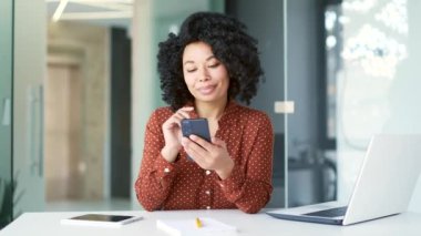 Genç Afro-Amerikan bayan çalışanı iş yerinde otururken akıllı telefon kullanıyor. Gülümseyen siyah kadın mesaj yazıyor, internette sohbet ediyor, sosyal medyada geziniyor ya da e-postaları kontrol ediyor.