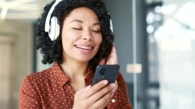 Ofiste oturmuş müzik dinleyen kulaklık takan mutlu Afro-Amerikan bayan çalışanı. Gülümseyen siyahi bir kadın, akıllı telefondan çalma listesini açar ve müzikten hoşlanır. Kapat.